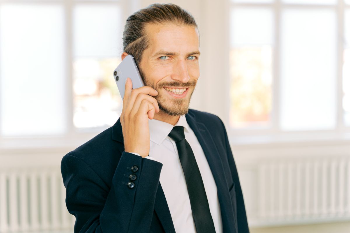 Smiling man in a dark suit and tie talking on a mobile phone beside a bright office window