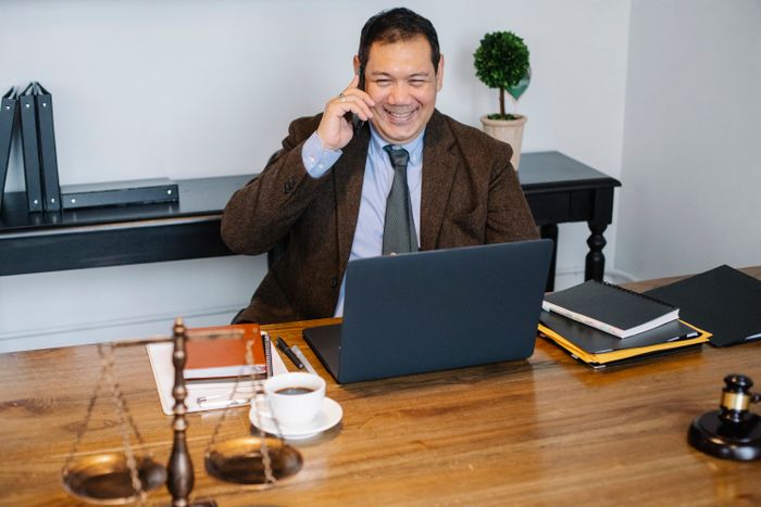 Joyful male lawyer wearing formal suit having conversation on mobile phone and using laptop while working in modern law firm