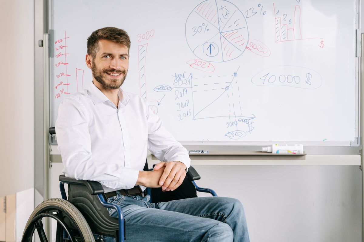 Smiling man in wheelchair at office, presenting business graphs on whiteboard
