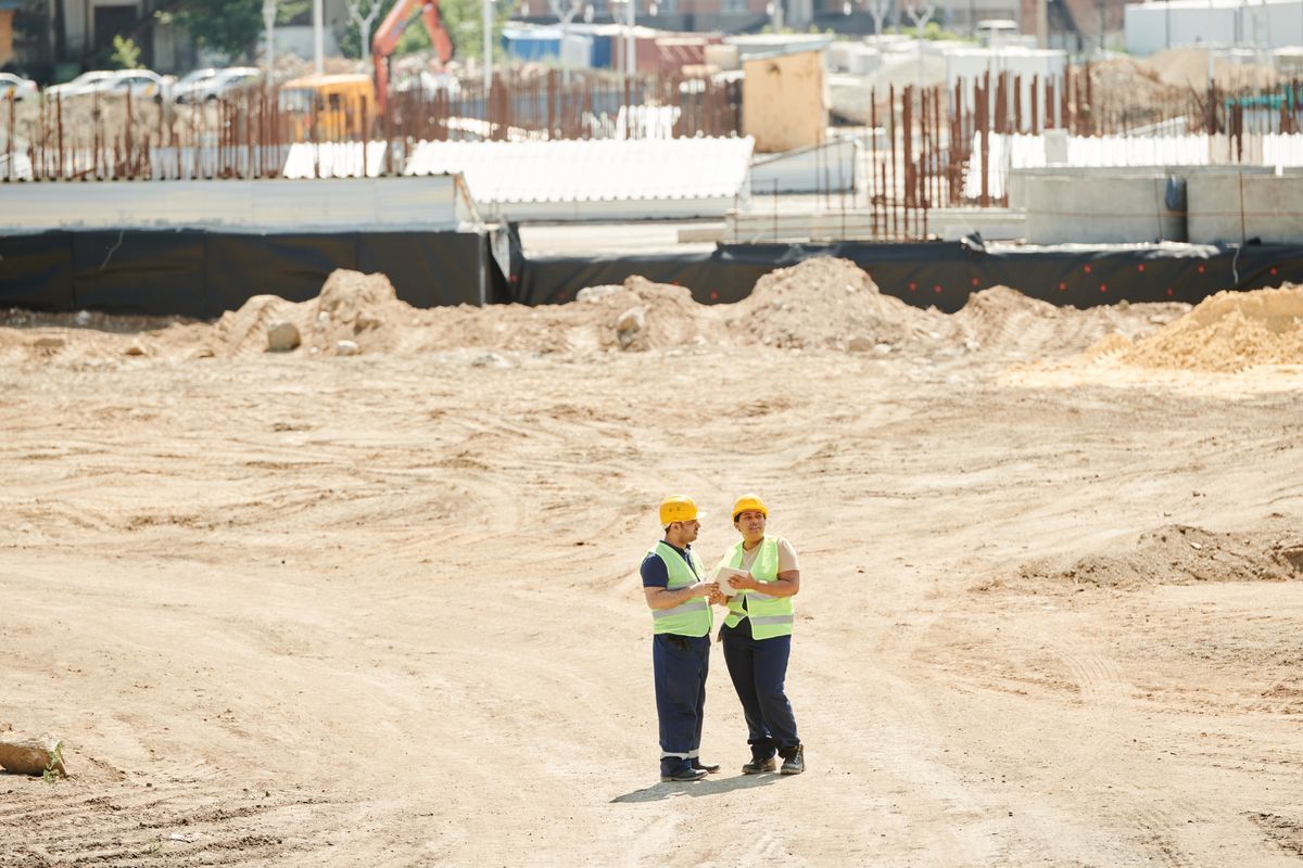 Two workers discussing plans on a sandy construction site