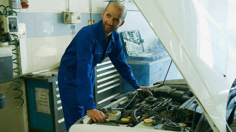 A smiling mechanic in a blue overall inspecting a car engine in a garage